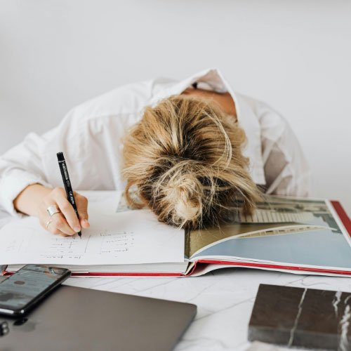Bild symbolisiert einen stillen Burnout und zeigt eine Frau die den Kopf auf dem Schreibtisch hat und weiterhin den Stift in der Hand hält, als wolle sie weiter arbeiten.